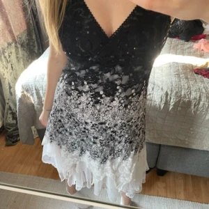 Svart och vit klänning med spetsdetaljer - Säljer en elegant svart och vit klänning med v-ringad urringning och spetsdetaljer. Klänningen har ett vackert blommönster och paljetter på överdelen. Den är ärmlös och har en volangkant nedtill.   Stl uk 14 motsvarar stl 38 