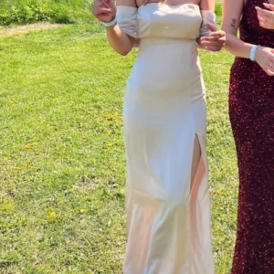 Champagnefärgad långklänning med slits - Säljer en elegant långklänning i champagnefärg med offshoulder-ärm och hög slits framtill. Klänningen har en figurnära passform och är tillverkad i ett glansigt satinliknande material. Perfekt till balen, älskar den men säljer då jag hittat en annan 💕(dragkedjan i ryggen är lite trög) säljs ej längre 