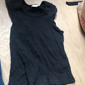 Svart ribbad topp med spetsdetaljer - Svart ribbad topp med korta ärmar och fina spetsdetaljer vid axlarna. Perfekt för en stilren och enkel look. Mjuk och stretchig, passar till mycket.