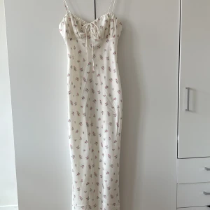 Vit långklänning med blommigt mönster och knyt - Superfin långklänning i vit med små rosa och gröna blommor. Smala axelband, söt knytning framtill och figurnära passform