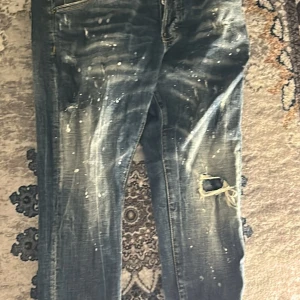 Dsquared jeans - Säljer ett par mörkblå jeans från Dsquared2 med coola slitningar och färgstänk. Jeansen har en trasig detalj vid knät och klassisk femficksdesign. Perfekta för dig som gillar en edgy streetstil.