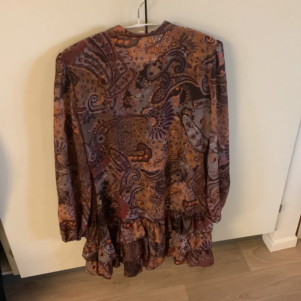 Säljer en unik klänning med volanger och knappar framtill. Klänningen har lång ärm och ett färgstarkt paisleymönster i lila, brunt och orange. Perfekt för dig som gillar bohemisk stil och vill sticka ut med en flowy känsla. Ha linne under. Inbyggd svart kjol. Neuletakit & Villapaidat.