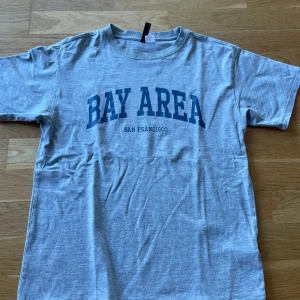 Grå t-shirt från H&M Divided - Säljer en grå t-shirt från H&M Divided med texten 'Bay Area San Francisco' i blått. T-shirten har en normal passform och är kortärmad. Perfekt för en avslappnad stil.