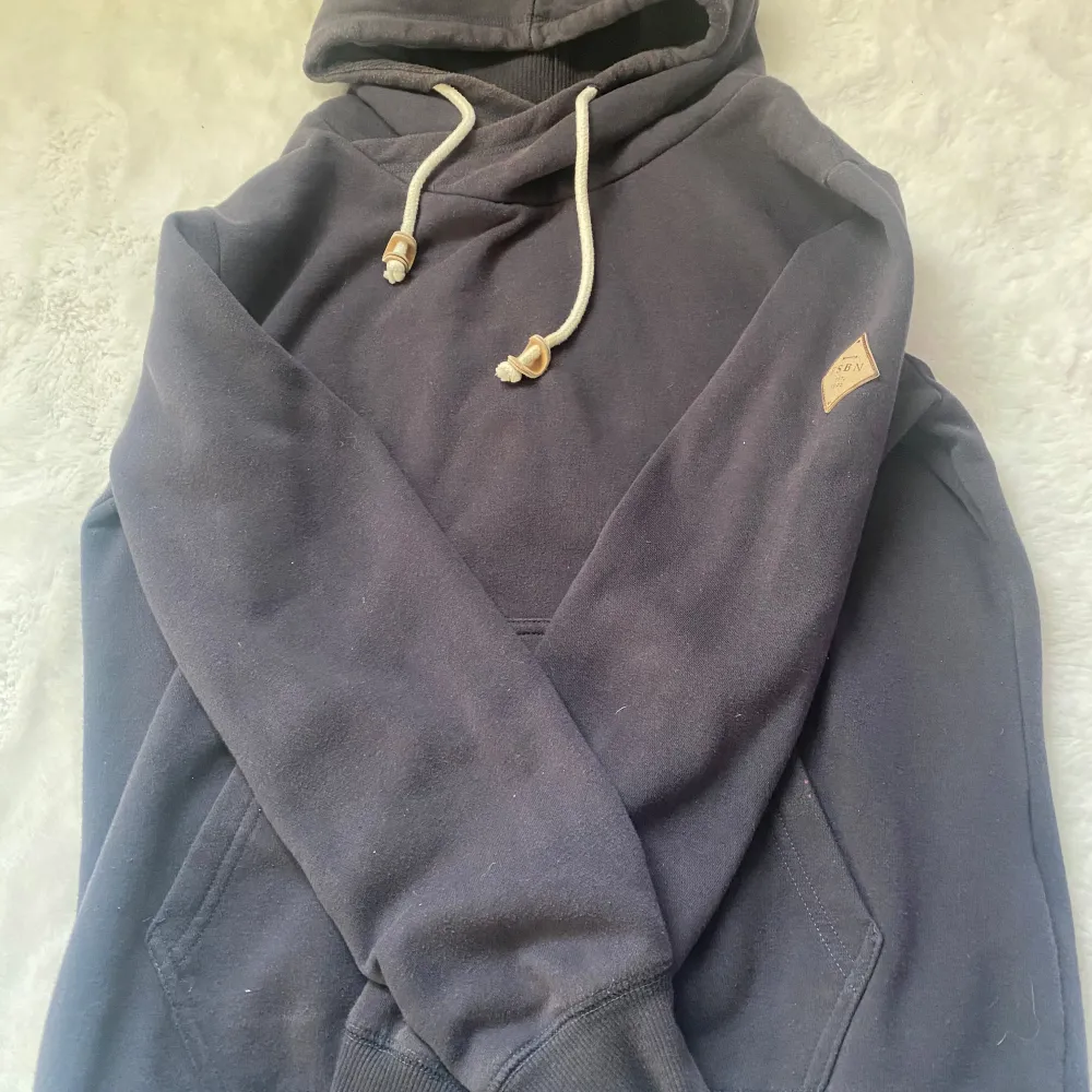 Säljer en stilren mörkgrå hoodie med snörning i huvan och en liten logga på bröstet. . Den har en klassisk känguruficka framtill.. Neuletakit & Villapaidat.