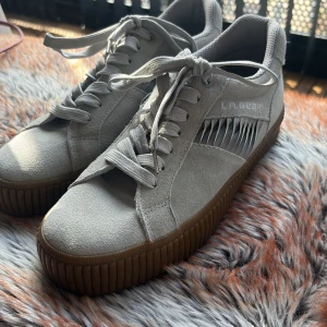 Grå sneakers från LA Gear - Snygga grå sneakers från LA Gear med snörning och en unik detalj på sidan. Skorna har en brun sula som ger en cool kontrast. Perfekta för en avslappnad stil. Aldrig använda 