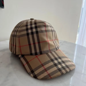 Rutig keps från Burberry - Snygg rutig keps från Burberry i klassiskt beige, svart och rött mönster. Kepsen har en justerbar rem baktill för perfekt passform och en broderad Burberry-logga. Perfekt för att ge din outfit en stilren touch. Äkta keps men utan låda osv där av pris