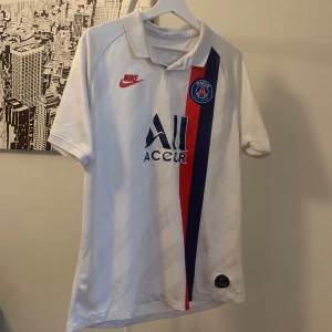 PSG vit bortatröja från Nike - Säljer en vit PSG fotbollströja från Nike med klubbmärke på bröstet och röd-blå diagonal rand. Tröjan har korta ärmar, klassisk krage och tryckt sponsorlogga på framsidan. Perfekt för dig som älskar fotboll och vill visa ditt stöd för Paris Saint-Germain.