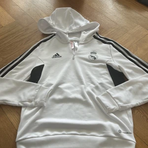 Vit Real Madrid hoodie från Adidas - Säljer en vit hoodie från Adidas med Real Madrid-logga på bröstet. Tröjan har svarta detaljer, dragkedja vid halsen och klassiska Adidas-ränder längs ärmarna. Perfekt för fotbollsfans att ha i vardagen eller när du tränar!