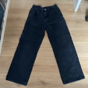 Svarta wide leg jeans från DAZY - Svarta jeans från DAZY i wide leg-modell med hög midja. Klassisk femficksdesign och raka ben. Jeansen är tillverkade i denim och har en enkel knapp- och dragkedjestängning framtill. De har ett håll i en av bakfickorna men inget som påverkar.