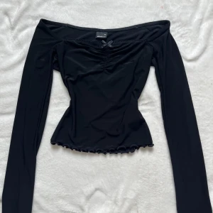 Svart långärmad topp med rosett  - Säljer en svart långärmad topp från Gina Tricot i storlek XS. Toppen har offshoulder-modell, liten rosett framtill och rynkdetalj vid bysten. Materialet är stretchigt och nederkanten har en vågig finish.