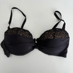 Svart spets-bh med dekorativa stenar - Snygg svart bh med spets upptill och små glittrande stenar. Kuporna är vadderade och har en dekorativ rosett och två små stenar i mitten. Axelbanden är justerbara. Använd. Osäker på storlek men gissar på 80D