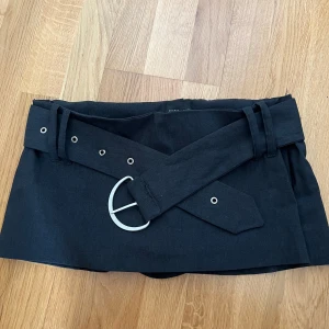 Svart minikjol med bälte - Säljer en svart kortkjol med tillhörande bälte och silverfärgat spänne. Kjolen har en stilren design med bred linning och bälteshällor. Perfekt för dig som vill ha en enkel men snygg kjol till garderoben.