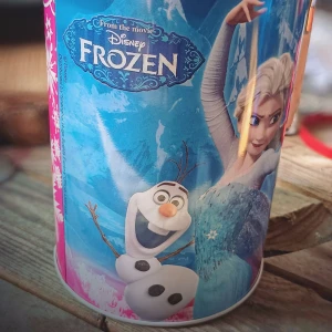 Frozen plåtburk med motiv - Säljer en plåtburk med motiv från Disneyfilmen Frozen. Burken har färgglada bilder av Elsa, Anna, Olaf och Sven mot en blå bakgrund med snöflingor. Perfekt som accessoar eller förvaring av småsaker.