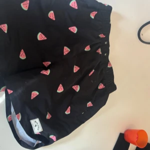 Svarta shorts med vattenmelonmönster - Snygga svarta shorts med ett lekfullt vattenmelonmönster. De har en elastisk midja och är fodrade med ett vitt nätmaterial för extra komfort. Perfekta för sommarens alla äventyr!