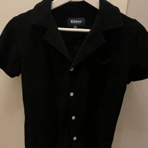 Svart kortärmad skjorta från Nikben - Snygg svart kortärmad skjorta från Nikben i en skön blandning av bomull och polyester. Skjortan har en klassisk krage och knappar framtill. Perfekt för en stilren look och skön till sommaren. Storlek 14 år! Nypris: 700kr
