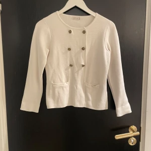 Vit kavaj från Ted Nicol - Elegant vit kavaj från Ted Nicol med dubbelknäppning och dekorativa knappar. Den har en klassisk design med långa ärmar och två fickor framtill. Perfekt för en stilren look.