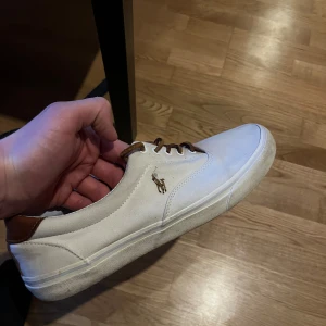 Vita Tygskor från Polo Ralph Lauren - Säljer ett par vita sneakers från Polo Ralph Lauren med bruna detaljer på häl och snörning. Skorna har rund tå och är tillverkade i textil med klassisk logga på sidan. Perfekta för en stilren och avslappnad look.