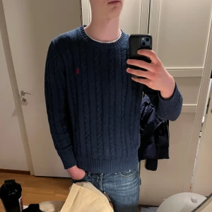 Mörkblå stickad tröja från Polo Ralph Lauren - Säljer en mörkblå stickad tröja från Polo Ralph Lauren med klassiskt kabelstickat mönster och rund hals. Tröjan har lång ärm och en liten röd logga broderad på bröstet. Perfekt för en stilren och avslappnad look.