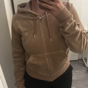 Beige velour hoodie från Juicy Couture - Säljer en beige velour hoodie från Juicy Couture med dragkedja och huva. Jackan har ribbade muddar, fickor framtill och broderad JC-logga på bröstet. Perfekt för en avslappnad och trendig look.