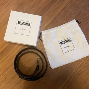 Svart och vit bälte från Moschino - Snyggt bälte från Moschino med svart och vit design. Bältet har en tydlig logotyp och ett stilrent spänne i silver. Perfekt för att ge din outfit en lyxig touch.