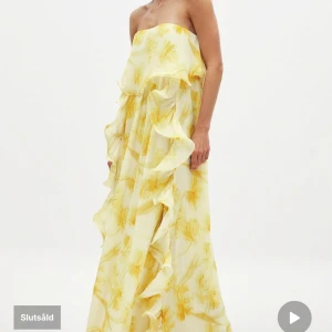 Gul axelbandslös långklänning NA-KD x Lojsan Wallin - Elegant gul axelbandslös långklänning med volanger och blommigt mönster. Slutsåld