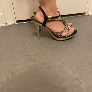 Svarta och guldiga klackskor från Buonarotti - Säljer ett par eleganta klackskor från Buonarotti med svarta remmar och glittriga detaljer i guld. Skorna har öppen tå, smala remmar med strass och hög klack. De är i ny skick i storlek 39-40! 