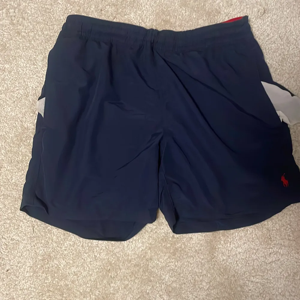 Snygga marinblå shorts från Polo Ralph Lauren med röd broderad logga. Perfekta för en stilren look. Tillverkade i ett lätt och bekvämt material med elastisk midja för extra komfort.. Shortsit.