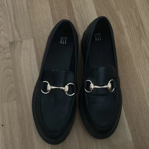 Svarta loafers från XIT - Snygga svarta loafers från XIT med dekorativt guldfärgat spänne på ovansidan. Perfekta för en stilren look.