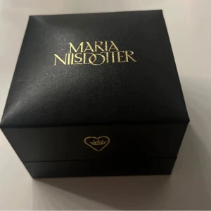 Smyckesask från Maria Nilsdotter - Elegant svart smyckesask från Maria Nilsdotter med guldfärgad logotyp på locket och ett hjärta med krona på framsidan. Perfekt för att förvara eller ge bort smycken.