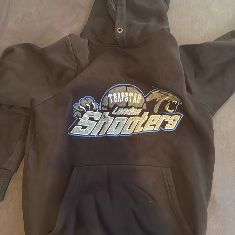 Säljer en svart hoodie från Trapstar med stort broderat London Shooters-motiv på bröstet i blått, vitt och gult. Hoodien har huva, magficka och ribbade muddar. Perfekt för dig som gillar streetwear och sportig stil.. Neuletakit & Villapaidat.