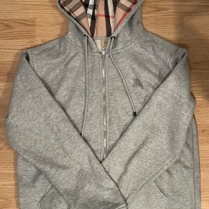 Grå hoodie från Burberry med dragkedja - Säljer en grå hoodie från Burberry i bomull med klassiskt rutigt foder i huvan och insidan. Tröjan har dragkedja, snörning i huvan och en broderad logga på bröstet. Perfekt för dig som vill ha en stilren och lyxig look.