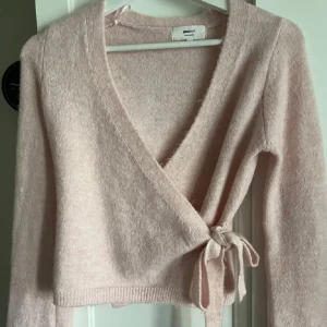 Ljusrosa omlottkofta från Gina Tricot - Supermysig ljusrosa omlottkofta från Gina Tricot med knyt i sidan och v-ringad hals. Koftan är stickad och har långa ärmar, perfekt för lager-på-lager under hösten och vintern. Snygg att matcha med jeans eller kjol för en chill look.