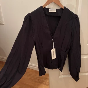 Svart blus från Carin Wester - Ny med prislapp!! En svart blus från Carin Wester med v-ringning och knappar framtill, uppknäppningsbar. Blusen har långa puffärmar och är tillverkad i ett glansigt, lätt skrynklat material. 