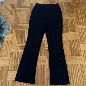 Svarta crepebyxor från NLY Trend - Svarta byxor från NLY Trend i crepematerial med hög midja och utsvängda ben. Snygg söm framtill och stilren look som passar till sneakers eller boots. Byxorna är i stretchigt polyestertyg och har en klassisk, clean vibe.