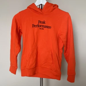 Orange hoodie från Peak Performance - Säljer en snygg orange hoodie från Peak Performance med svart tryck på bröstet. Tröjan har huva, magficka och långa ärmar. Perfekt för dig som gillar sportig och avslappnad stil. Materialet är mjuk bomull som känns skönt mot huden.