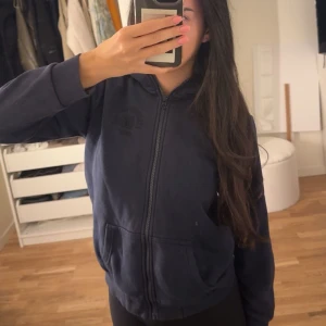 Mörkblå kofta Peak Performance - Mörkblå hoodie från Peak Performance med broderad logga och siffran 86 på bröstet. Tröjan har dragkedja, huva och två fickor framtill. Perfekt för dig som gillar en sportig och avslappnad stil.