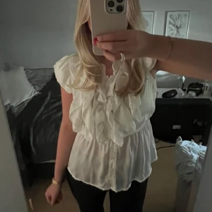Vit blus med volanger och knappar - Supersöt vit blus med volanger framtill och knäppning hela vägen ner. Blusen är ärmlös och har en lätt markerad midja med resår 🩷