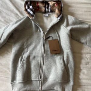 Grå hoodie från Burberry med dragkedja - Snygg grå hoodie från Burberry med klassiskt rutigt foder i huvan. Tröjan har dragkedja framtill, fickor och snörning vid huvan. Perfekt för dig som gillar stilrena och exklusiva plagg med en twist av ikoniskt mönster.
