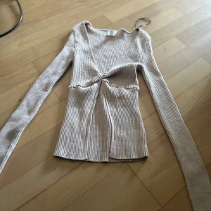 Beige ribbad omlottopp med v-ringning - Säljer en beige ribbad omlottopp med lång ärm och snygg v-ringning. Toppen har en twistad detalj framtill som ger en cool look och är figurnära i passformen. Perfekt att styla med jeans eller kjol för en trendig outfit.