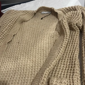 Beige stickad kofta med grov struktur - Säljer en beige stickad kofta från 157 med grov och mysig struktur. Koftan har öppen front och ribbade kanter, perfekt att slänga på sig när det är lite kyligt. Passar dig som gillar en chill och enkel stil. Storlek 160.