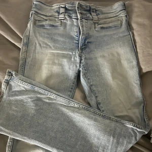 Ljusblå bootcut jeans med dubbla knappar från h&m - Säljer ett par jättefina ljusblå jeans med bootcut-snitt och dubbla knappar i midjan. Jeansen har klassiska bälteshällor och fickor fram, samt en snygg tvättad look. Perfekta för dig som gillar en avslappnad men trendig stil.