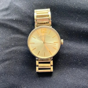 Guldig klocka Marc by Marc Jacobs - Stilren guldig klocka från Marc by Marc Jacobs med rund urtavla och länkarmband i rostfritt stål. Klockan har minimalistisk design med tydliga index och stor 12:a upptill. Perfekt accessoar för att lyfta din outfit.        Den har fått lite färg på baksidan och armbands uret är 34 mm. Den behöver ett nytt batteri.