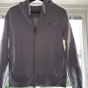 Grå hoodie med dragkedja från Morris - Snygg grå hoodie från Morris med hel dragkedja, huva med vita snören och broderad logga på bröstet. Tröjan har ribbade muddar och två fickor framtill. Perfekt för en chill och stilren look. Passar dig mellan 163-170 ca. Tröjan är inte jätte använd och är i ett mycket bra skick.