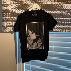 Svart t-shirt L'ARTE med tryck - Svart t-shirt från L'ARTE, tillverkad i kraftig bomull. Framsidan har ett stort svartvitt tryck av en person med solglasögon som röker. Klassisk passform och rund hals.