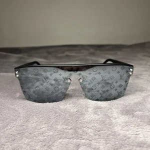 Lv solglasögon  - Snygga svarta solglasögon från Louis Vuitton med spegellinser i shield-modell. Linserna har LV:s ikoniska monogrammönster och bågarna är mattsvarta med diskret logga på sidan. Perfekt accessoar för dig som vill sticka ut med lyxig streetstyle.