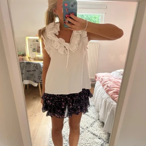 Vit volangblus - Supersöt vit blus med stora volanger runt v-ringningen och ett knyt framtill. Ärmlös och luftig modell som ger en romantisk vibe. Perfekt till kjol eller jeans för en trendig look.