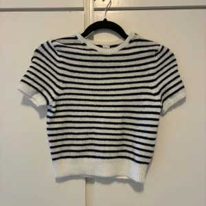 Randig stickad kortärmad tröja - Säljer en mysig stickad tröja med svarta och vita horisontella ränder. Tröjan har korta ärmar, rund hals och ribbad kant vid midjan och ärmslut. Perfekt för dig som gillar klassisk stil med en modern twist.