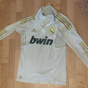 Real Madrid långärmad Ronaldo-tröja - Säljer en långärmad Real Madrid fotbollströja från Adidas med Ronaldo och nummer 9 på ryggen. Tröjan är vit med guldiga detaljer och Adidas klassiska ränder på ärmarna. Tillverkad i lätt och ventilerande material (Climacool) och har klubbmärket på bröstet.