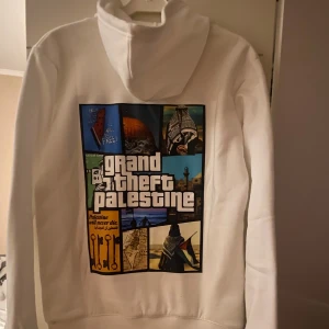 Vit hoodie Grand Theft Palestine - Vit hoodie med svart tryck 'grand theft palestine' på framsidan och stort färgglatt motiv på ryggen med collage och text. Klassisk känguruficka, huva med snörning och mjukt material. 