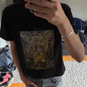 Svart t-shirt med konsttryck från La Vichy - Svart t-shirt från Le Vichy med stort färgstarkt konsttryck på bröstet och texten 'VICTORY' ovanför motivet. Klassisk passform med korta ärmar och rund hals. Liten broderad patch på ärmen. Perfekt för dig som gillar unika prints och streetstyle.